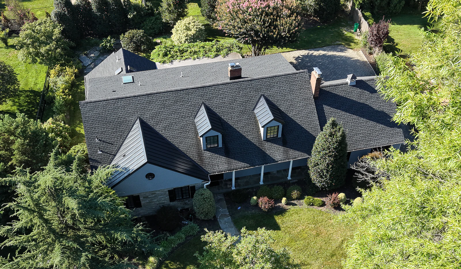 Aerial view of a large slate-roof colonial home with dormer windows, covered porch and mature landscaping